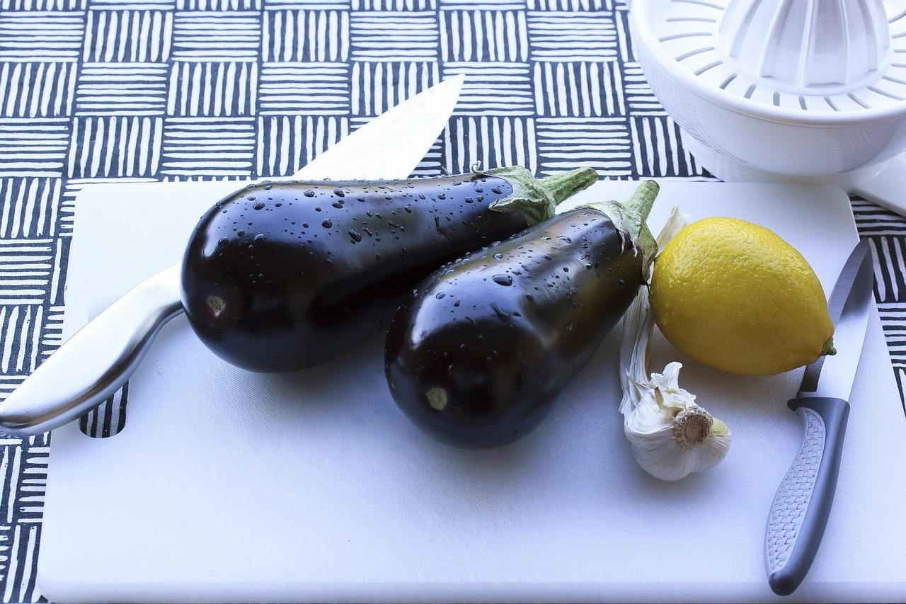 Zwei Auberginen, eine Zitrone und Knoblauch liegen auf einem weißen Schneidebrett aus Plastik, daneben zwei Küchenmesser