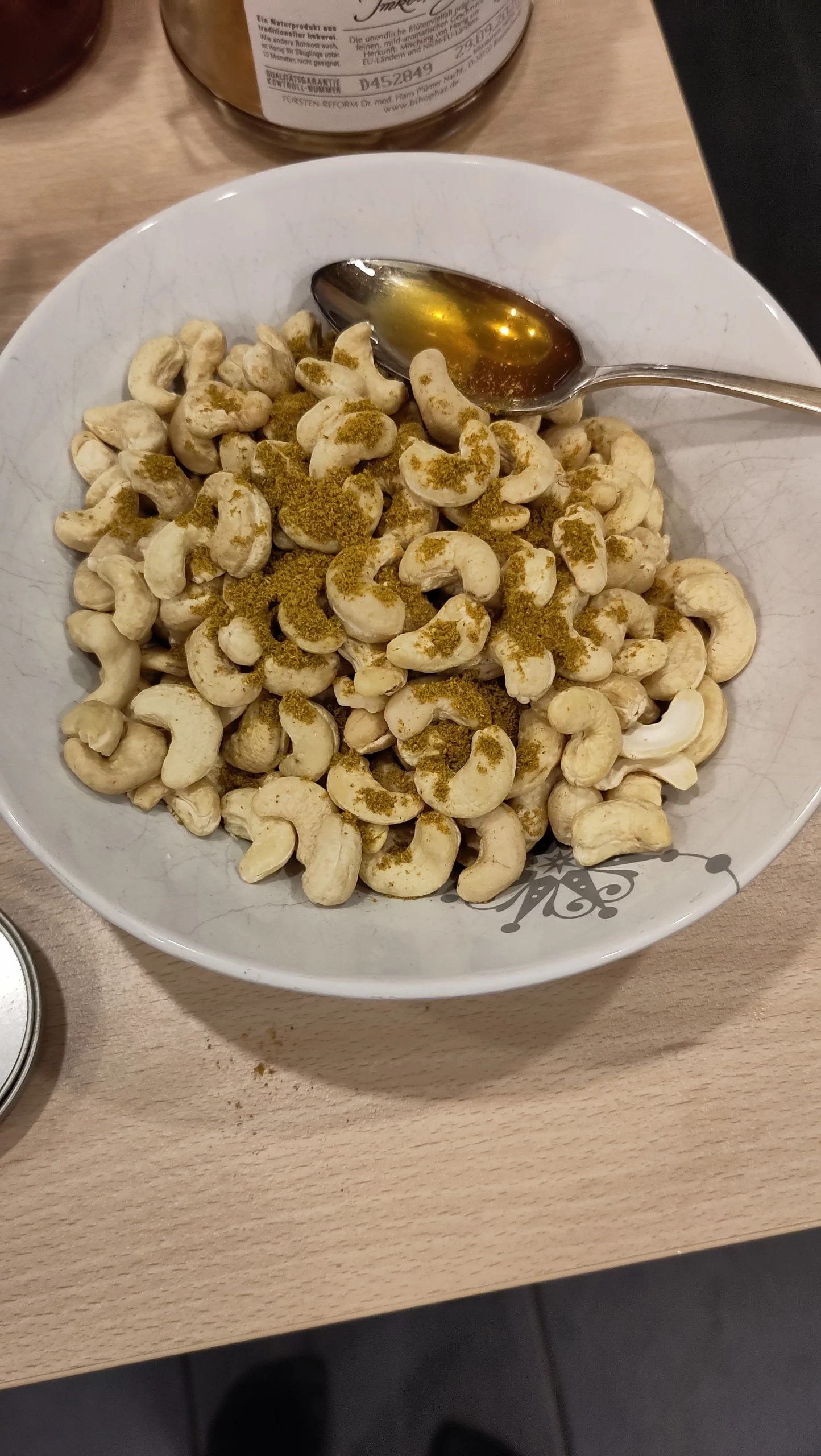 Das Bild zeigt eine Schale mit Cashews, die mit Currypulver bestreut sind und einen Löffel Honig.
