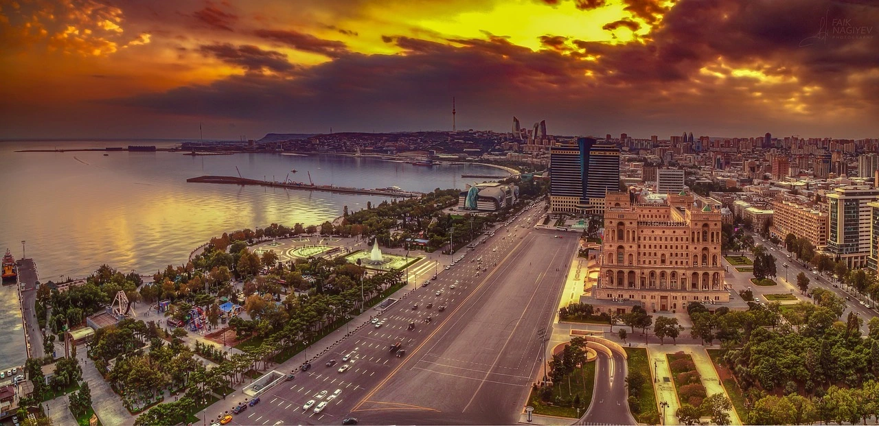 Das Foto zeigt die Luftaufnahme der Stadt Baku. Zu sehen ist ein Stück Meer, eine mehrspurite Straße und einige Gebäude. Der Himmel ist rötlich gefärbt vom Sonnenuntergang.