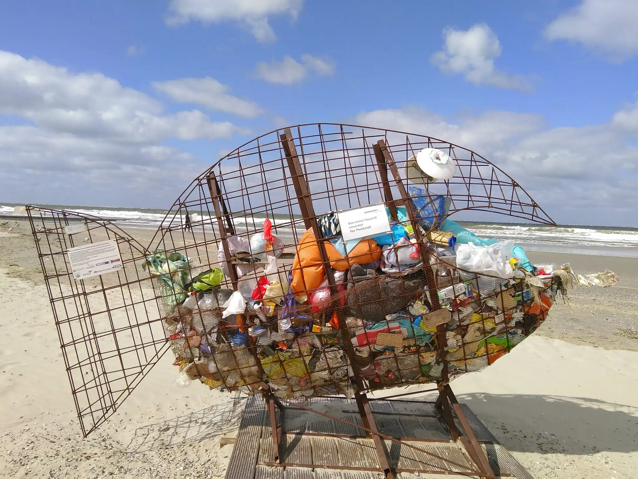 Am Strand steht ein großer Müllkorb in Form eines Fisches aus einem Drahtgestell, sein "Mageninhalt" sind Plastiktüten.