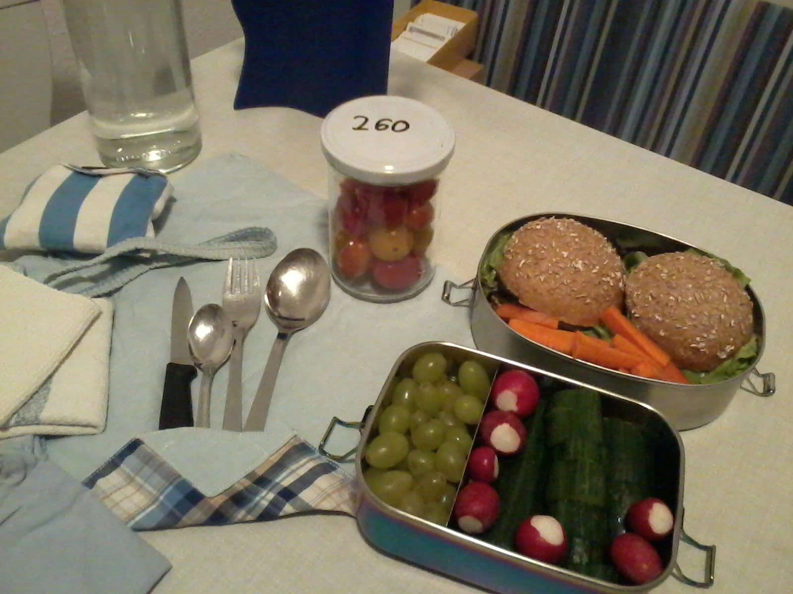 Das Foto zeigt eine gefüllte Glasflasche mit Wasser, eine Edelstahldose mit Brötchen, Möhrensticks, Trauben und Gurke, ein Glas mit Tomaten sowie ein Besteckset.