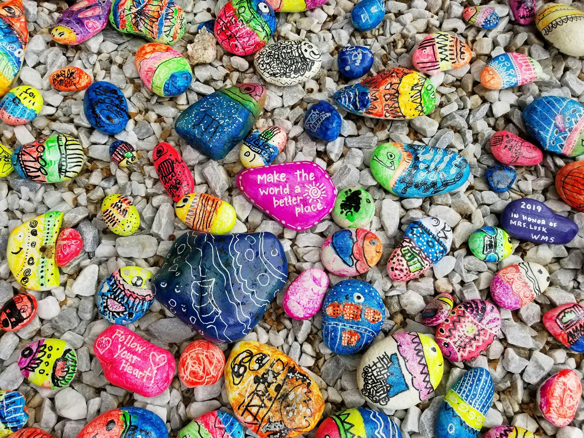 Viele bunte Steine liegen auf grauem Kies. Manche sind mit motivierenden Botschaften beschriftet, zum Beispiel: "Make the World a better place" oder "Follow your Heart!"