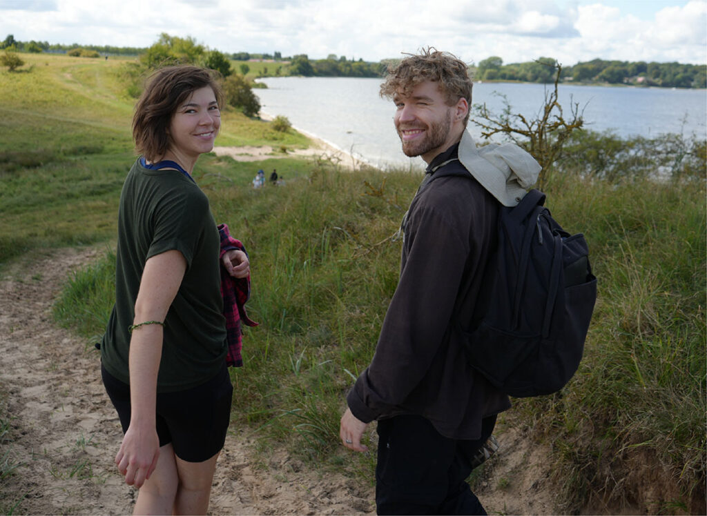 Silka und Marcel drehen sich beim Gehen entlang der Ostseeküste um und lächeln in die Kamera.
