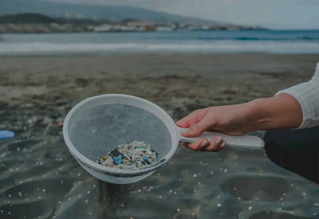 Eine Hand hält ein Sieb, dass Müll aus feinem Sand siebt.
