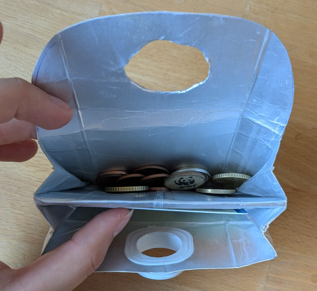 Das fertige Portemonnaie wird mit einer Hand offen gehalten. Darin ist in dem hinteren Kompartiment Kleingeld und im vorderen eine Karte zu sehen.